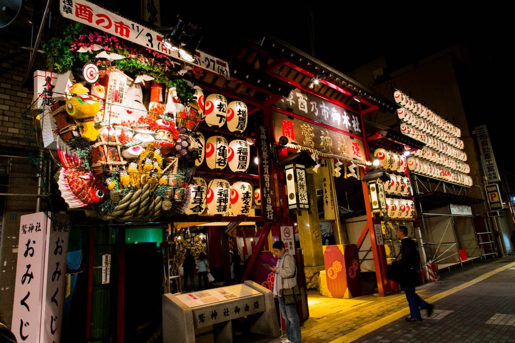 夜の鷲神社の入口。赤い門の上に「鷲神社」の額が掛かり、左に松や俵、鯛を組み合わせた大きな飾り熊手、右に奉納の提灯が並ぶ