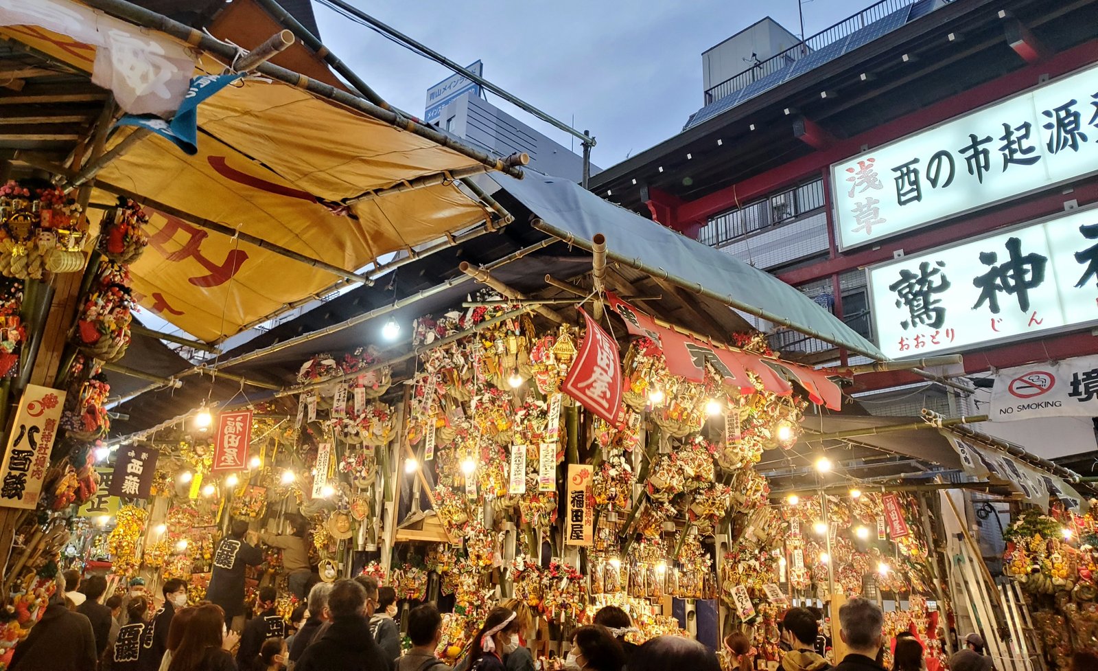 夕暮れの浅草の酉の市。飾り熊手をぎっしり掛けた露店の屋根越しに、「酉の市起源発祥 鷲神社」の白い看板が見える