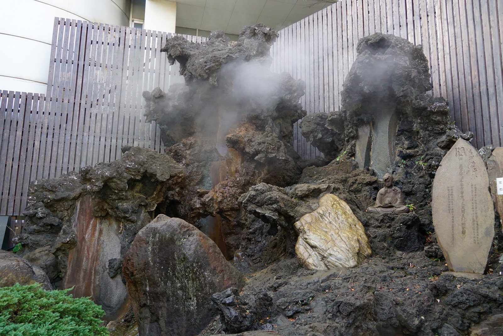 熱海の大湯。黒い溶岩のような岩の間から湯けむりが立ちのぼり、右手に座った石仏と文字を刻んだ石碑があり、背後を木の板塀が囲んでいる
