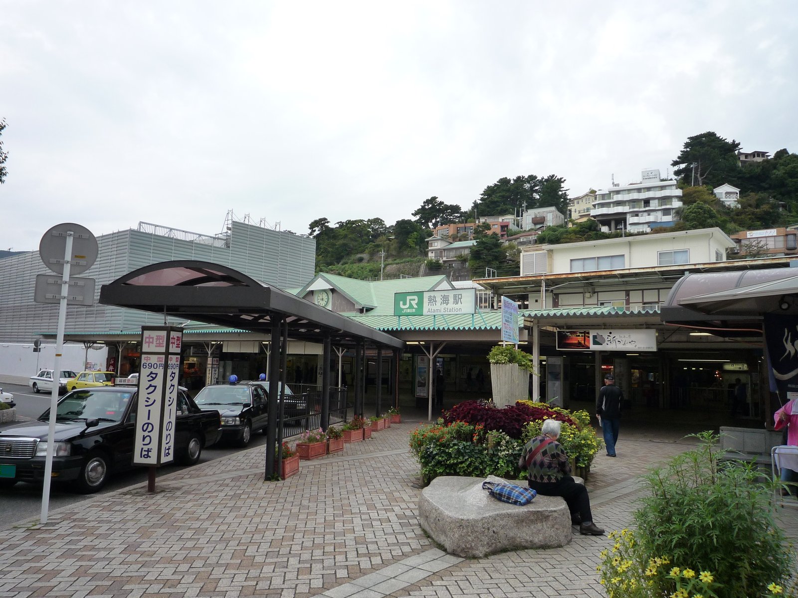 JR熱海駅の駅舎。緑の屋根に「JR 熱海駅 Atami Station」の看板があり、手前の駅前広場に花壇とタクシーのりば、背後の山の斜面に建物が見える