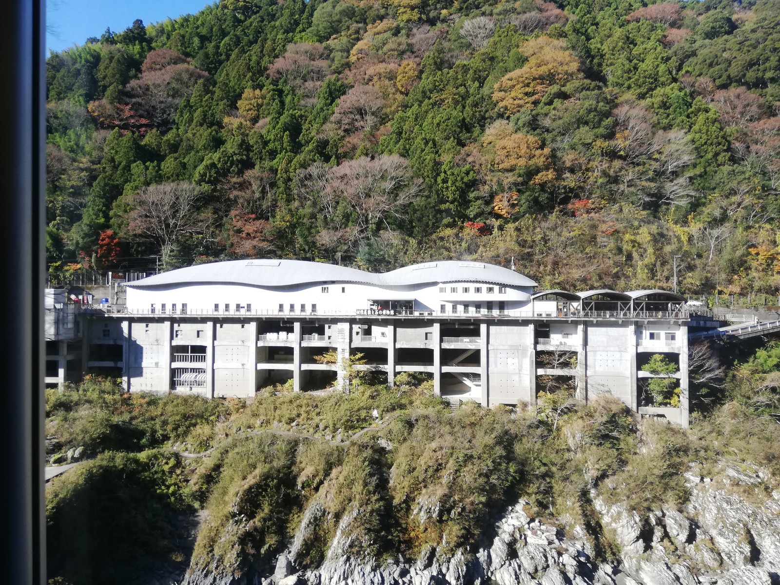 吉野川の深い渓谷にせり出すように建つ道の駅大歩危の建物。手前は切り立った岩肌の渓谷で、奥の山は紅葉している。屋上に「妖怪屋敷と石の博物館」の看板が見える