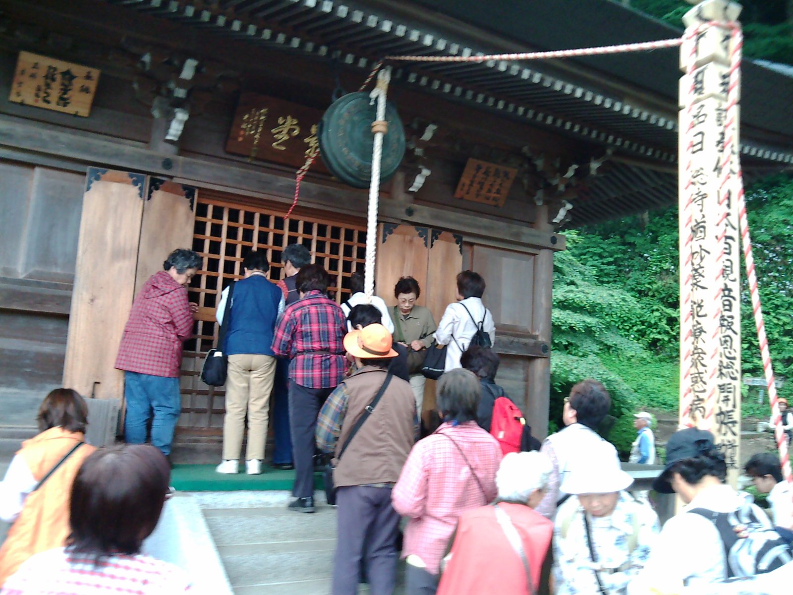 秩父札所27番 大渕寺のお堂の前に集まった参拝者。格子戸の堂の軒に鰐口が下がり、右手に「日本百観音報恩総開帳」と書かれた柱が立つ
