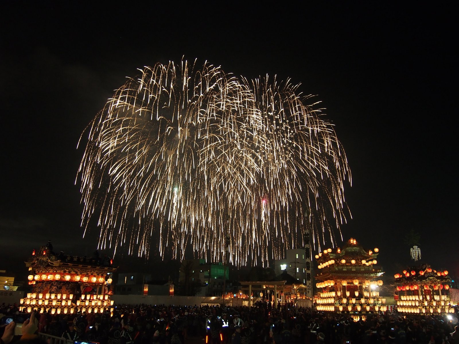夜空に大きく開いた花火の下に、提灯を一面に灯した山車が 4 基並び、手前に見物の人の群れ