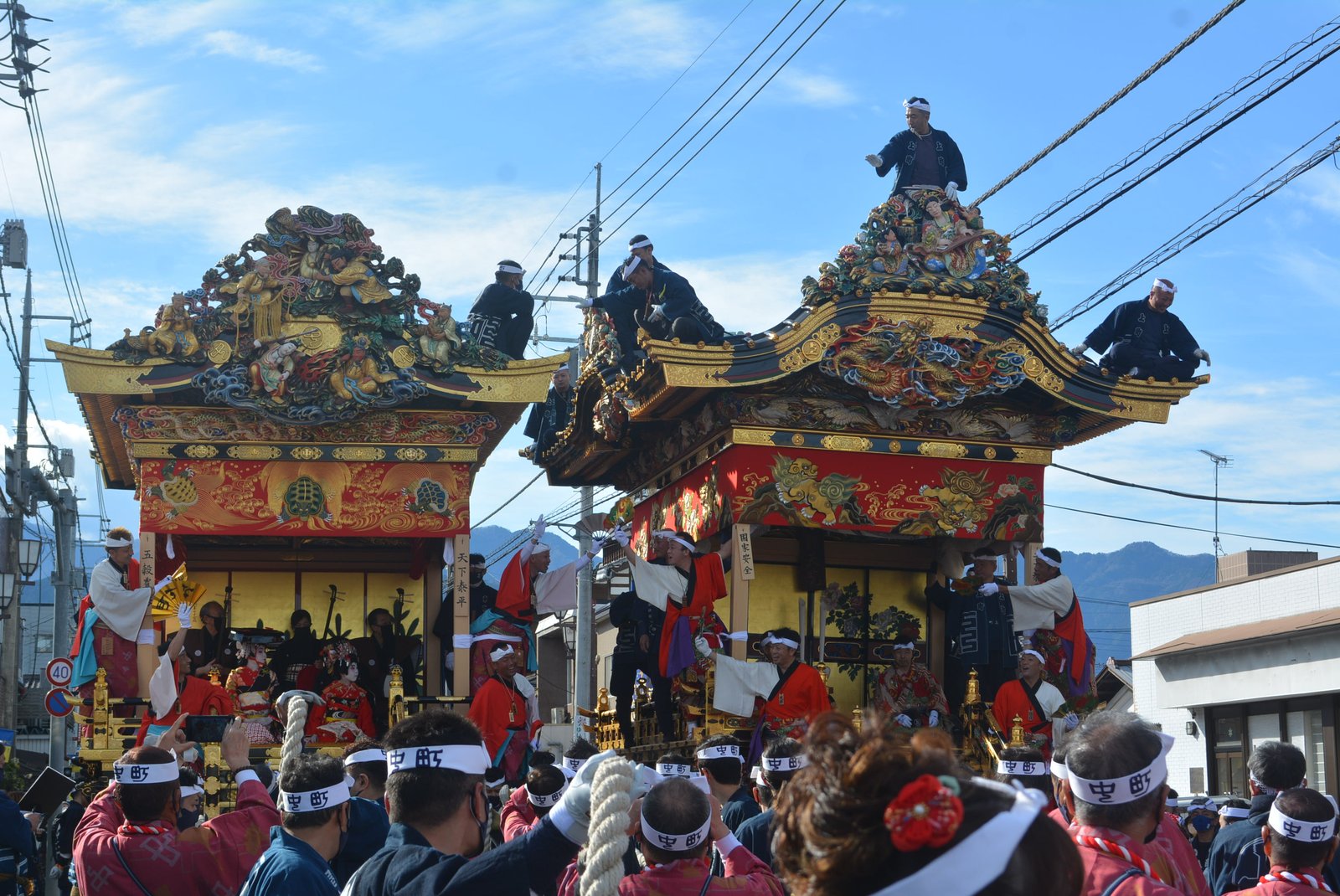 昼の秩父の町なかで、金色の彫刻と赤い幕で飾られた屋台 2 基が道ですれ違う。屋根の上に法被の人が乗り、手前に「中町」の鉢巻きをした曳き手が集まっている