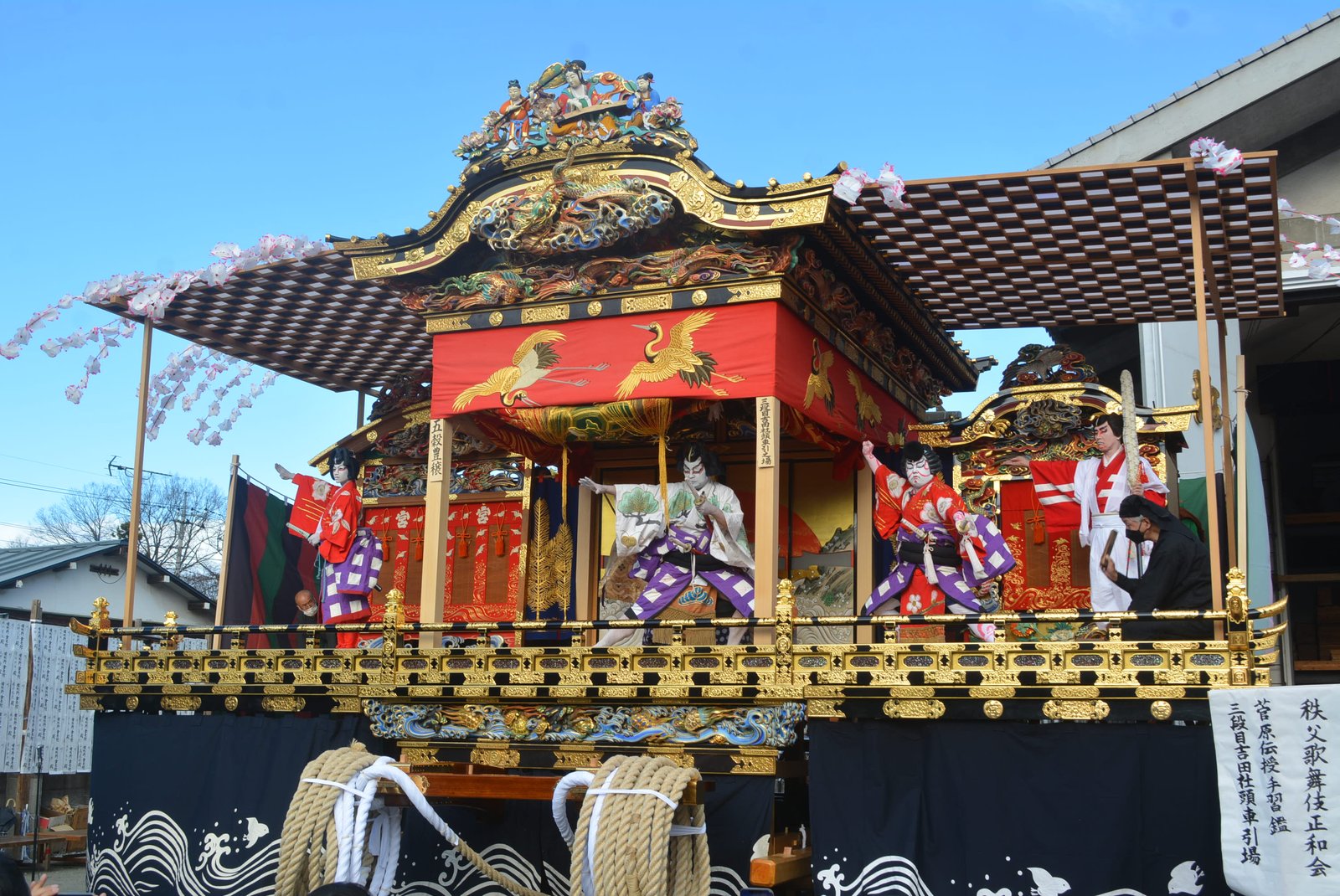 屋台の左右に張り出した舞台で歌舞伎が演じられている。鶴の模様の赤い幕と金色の彫刻の屋根の下に、隈取りをした役者が 3 人立ち、脇に「秩父歌舞伎正和会」の幟