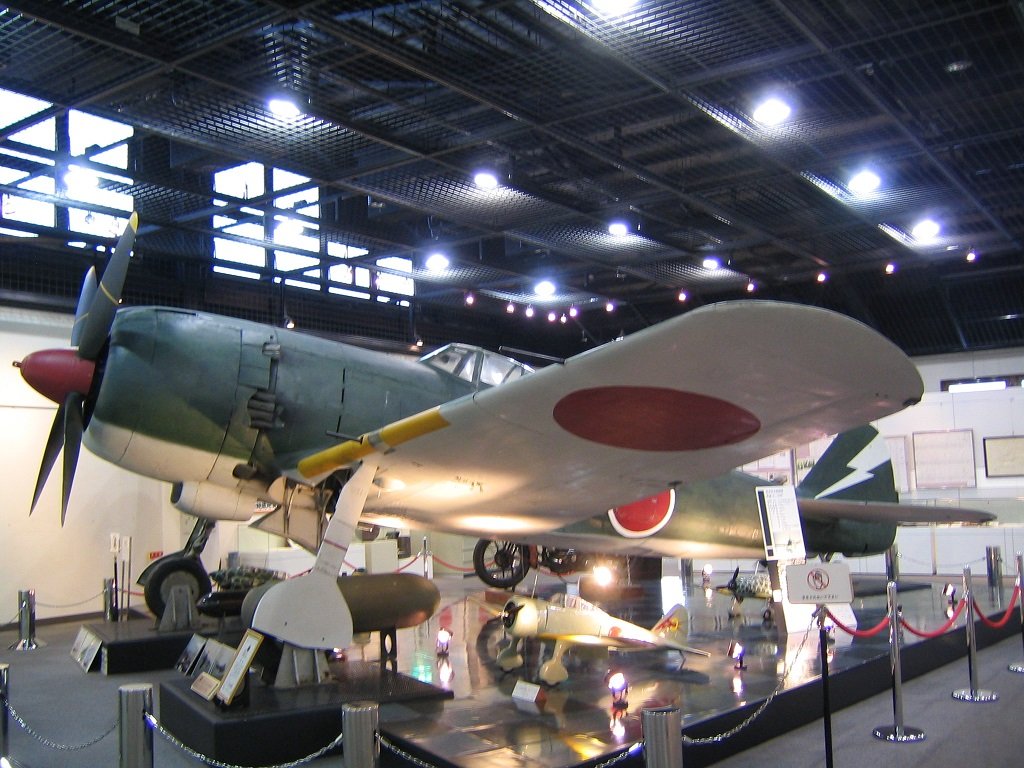 展示室に置かれた緑色の四式戦闘機。主翼に赤い日の丸が付き、左に4枚羽根のプロペラと赤いスピンナーがあり、手前に小さな機体の模型や解説板が並ぶ