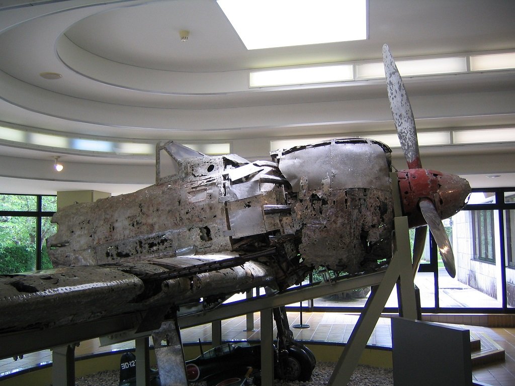 円形の天井の展示室に置かれた、表面が腐食して白茶けた零戦の機体。胴体とエンジン部分の金属板に穴や傷みが多く、先端に赤いプロペラの軸と1枚のプロペラが残る