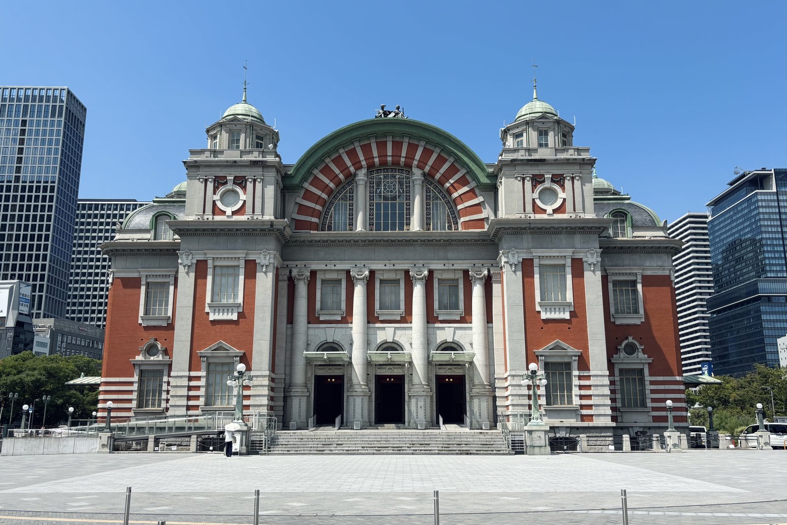 青空の下、赤レンガと白い石の柱、緑青の屋根を持つ大阪市中央公会堂の正面。手前に石畳の広場と3つの入口がある