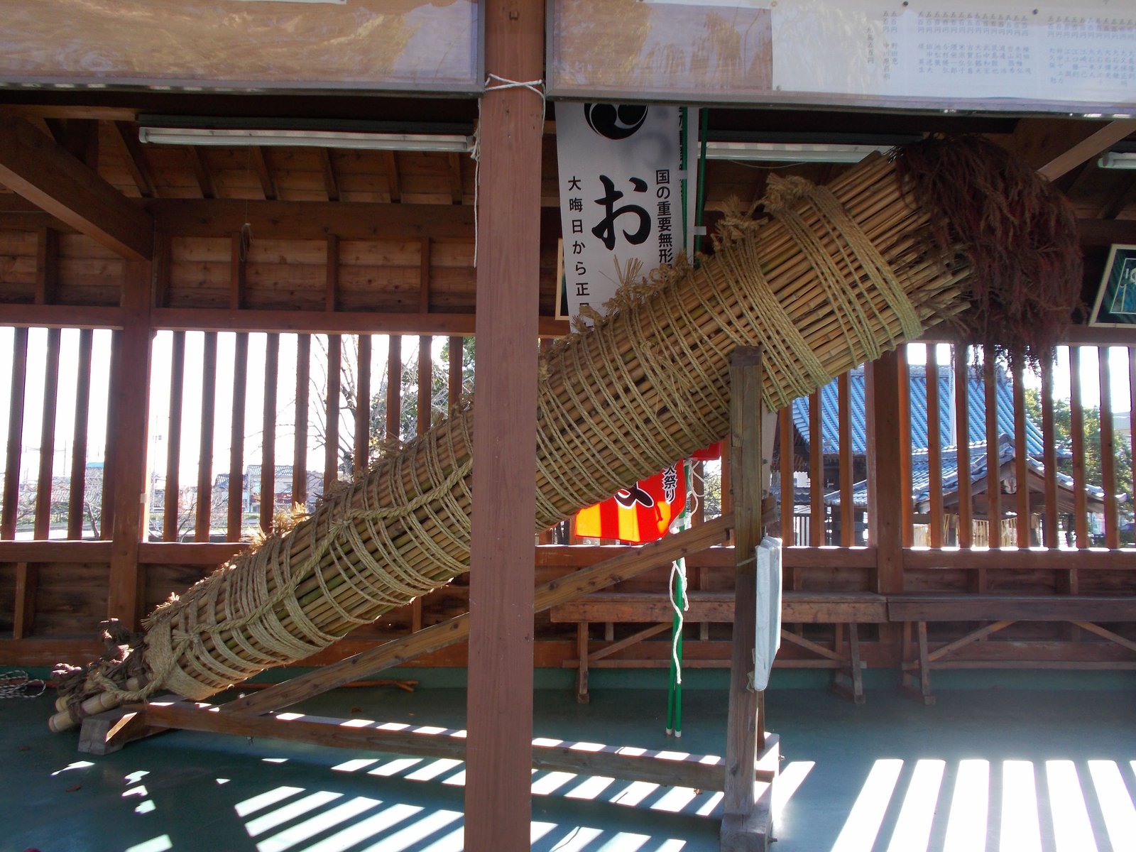 柱と腰壁だけの小屋の中に、斜めに据えられた大松明。細い茎を縄で幾重にも巻いて太く束ねたもので、先端は赤茶色にほぐれている。後ろののぼりには「国の重要無形」「大晦日から正」の文字