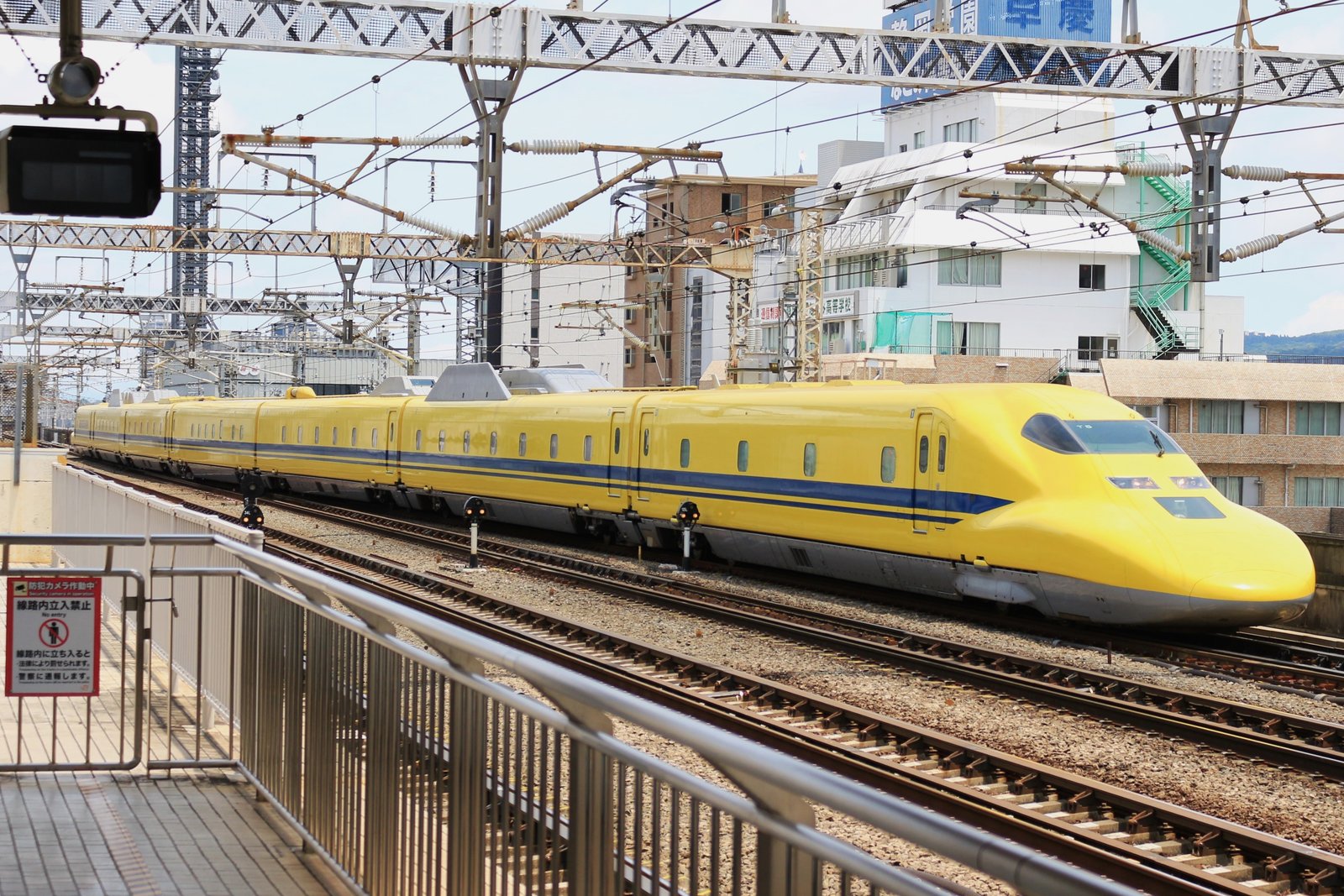 駅のホームの脇を通過する黄色い車体に青い帯の7両編成のドクターイエロー。背後にビルと架線柱が並ぶ