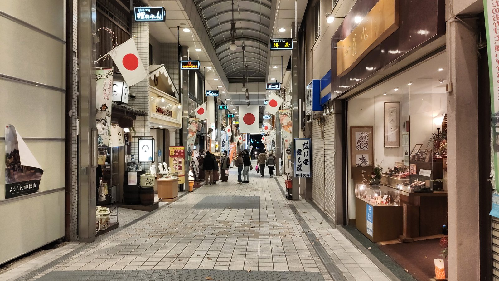 アーケードの商店街。天井から日の丸の旗が並んで下がり、両側に土産物や菓子の店が明かりをともして並び、タイル敷きの通路の奥を人が歩いている