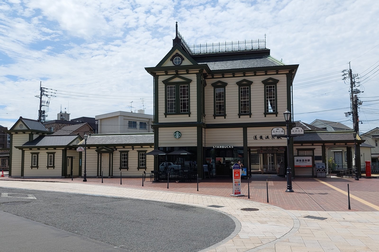 クリーム色の板張りの洋風駅舎。中央に緑の窓枠の2階建て、屋根に鉄の飾り柵があり、1階に道後温泉駅の看板とカフェ、手前にレンガ敷きの駅前広場が広がる