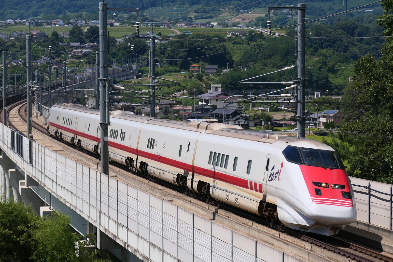 高架橋のカーブを走る白い車体に赤い帯の6両編成の検測車。先頭車の側面に「East i」のロゴ、背後に住宅と山