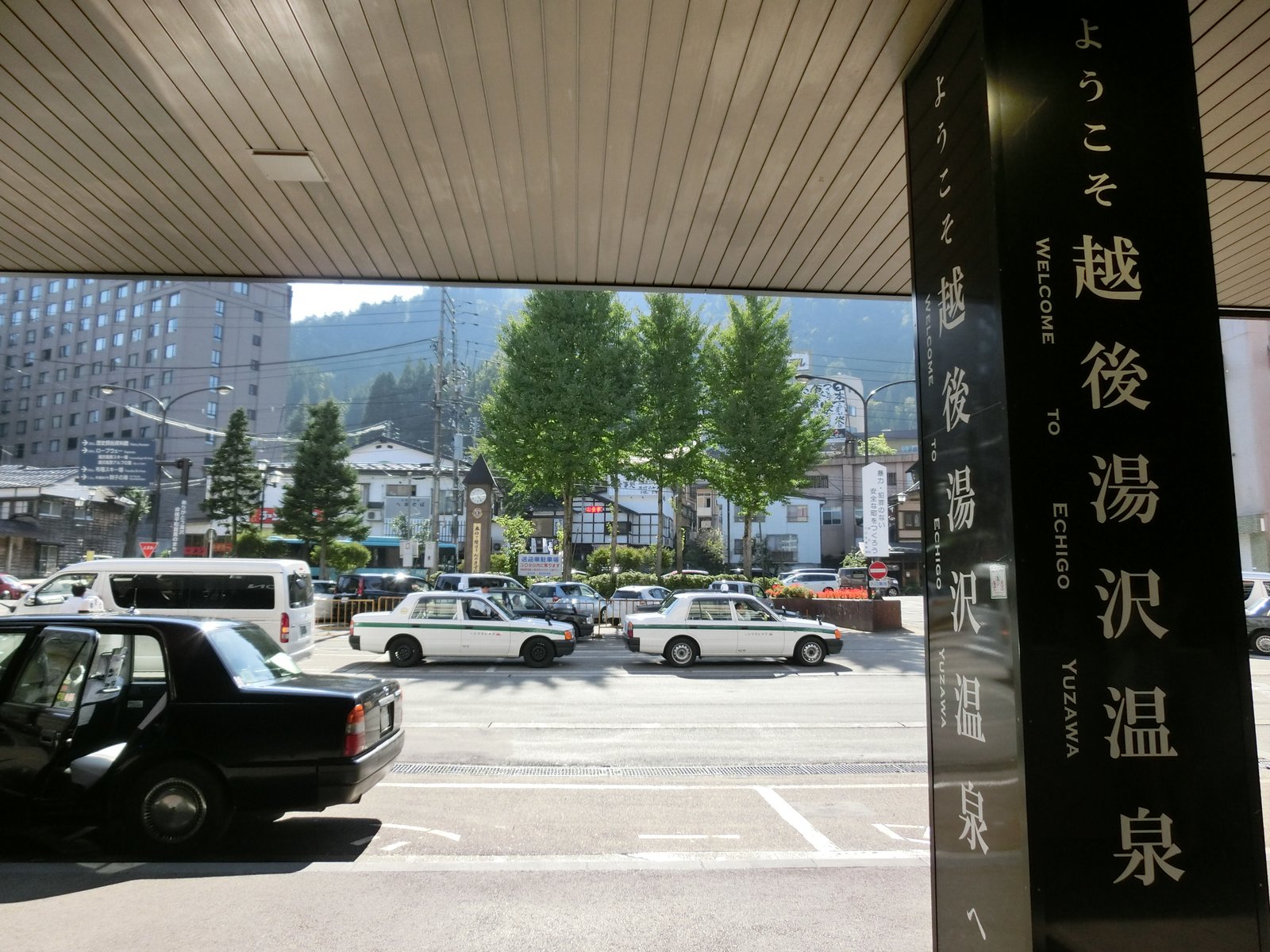 越後湯沢駅の屋根の下に立つ黒い柱に「ようこそ越後湯沢温泉へ」の白い文字。その向こうにタクシーが並ぶ駅前の通りと、木々の奥の建物、背後の山が見える