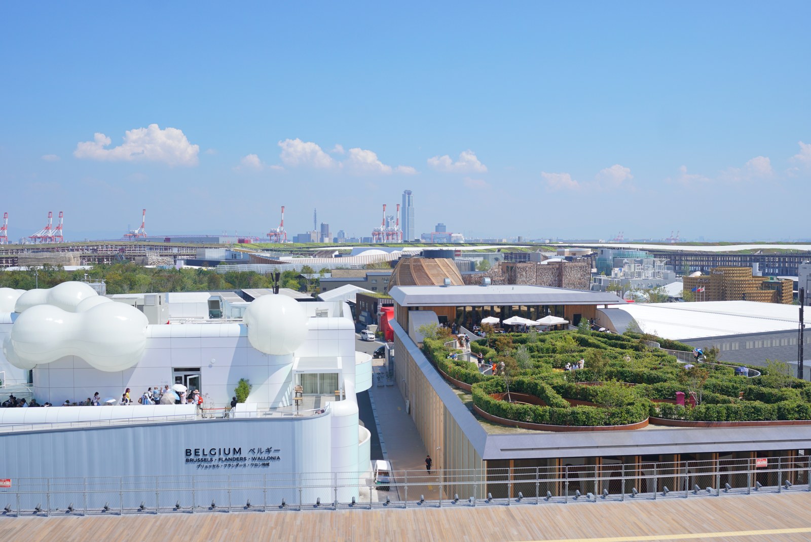 大阪・関西万博の会場風景。ベルギー館の白い外観の奥に緑の屋上庭園と港のクレーン、大阪の街並みが見える