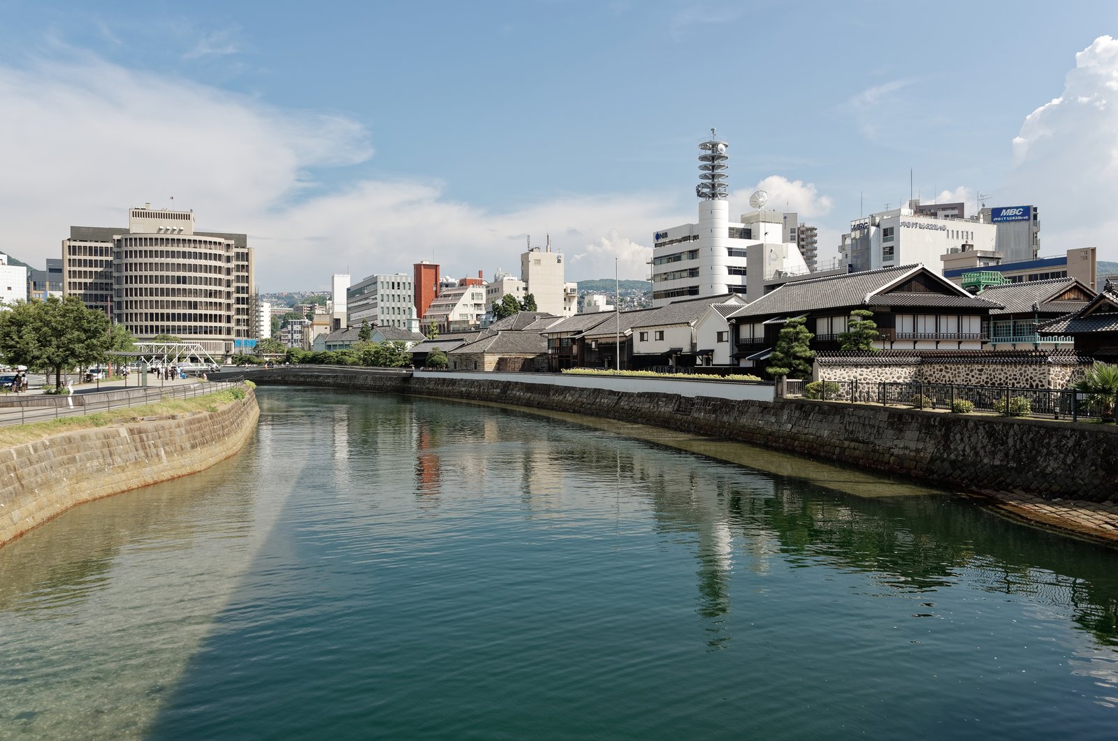 長崎の出島。水路の石積みの岸の上に、白い漆喰の壁と瓦屋根の建物が並び、奥に現代のビルが立つ