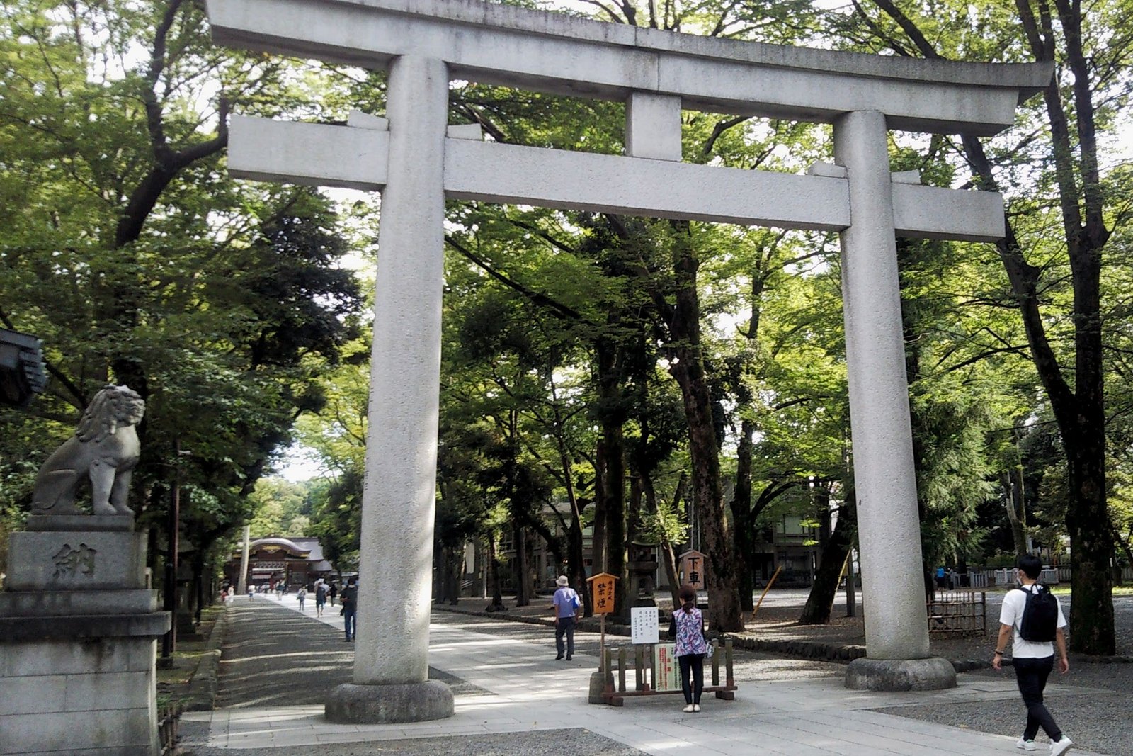 大國魂神社の大鳥居。木立の間の参道の奥に随神門が見え、左に狛犬、参拝者が歩いている