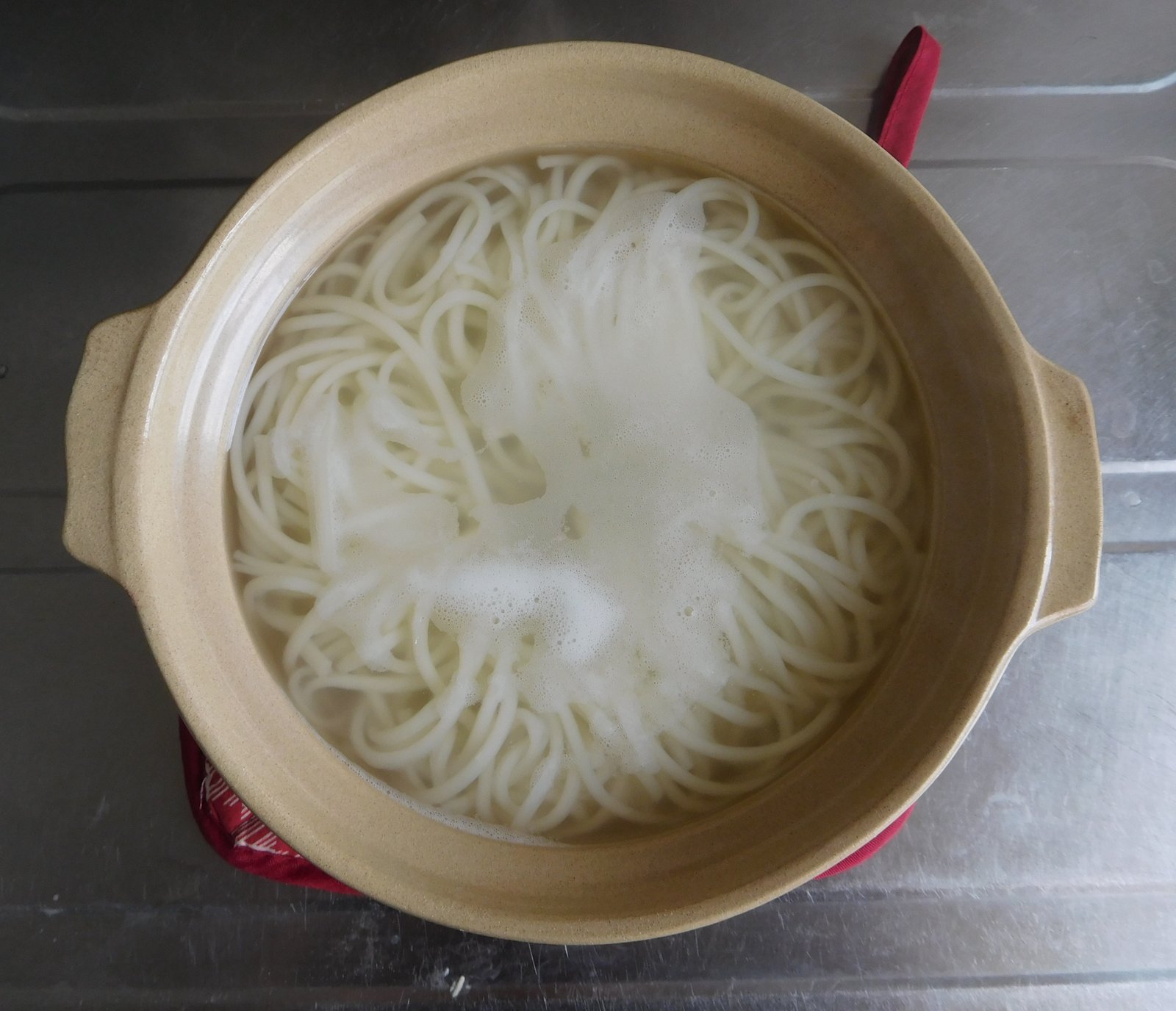 赤い鍋敷きの上に置いた土鍋の写真。湯の中で白く細いうどんが渦を巻くようにゆだち、真ん中に泡が浮いている