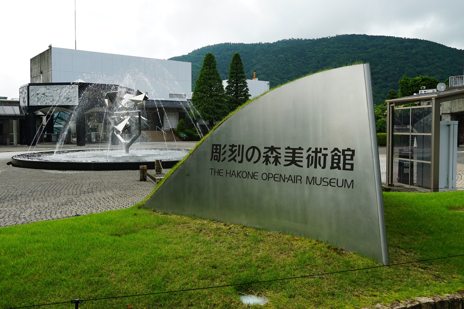 彫刻の森美術館の入口に立つ銀色の館名サイン「彫刻の森美術館 THE HAKONE OPEN-AIR MUSEUM」。奥に噴水と本館の建物、背後に緑の山が見える