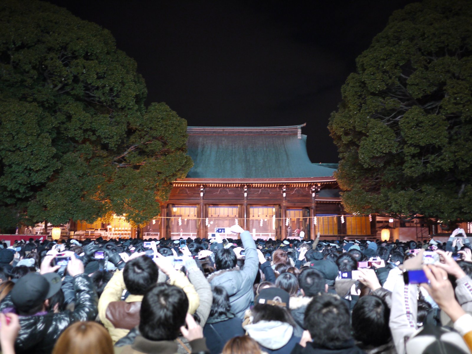夜の明治神宮の社殿の前を埋める大勢の参拝者。ほとんどが後ろ姿で、掲げた携帯電話の画面が光っている。奥に緑の屋根の社殿、両脇に大きな木