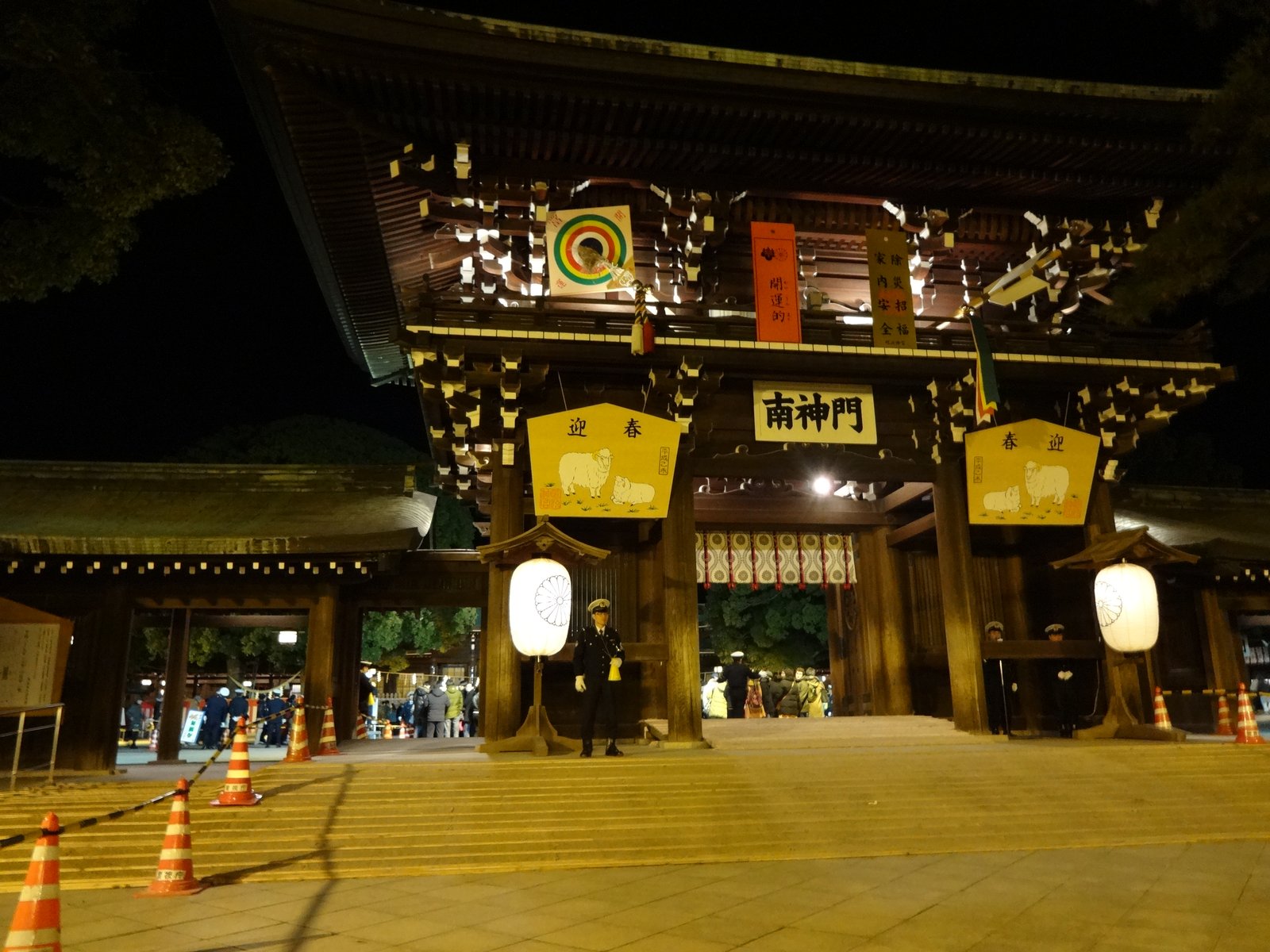 夜の明治神宮の南神門。門に迎春の絵馬と白い提灯が掛かり、石段の下に赤白のコーンが並ぶ。門の前に警備の人が立ち、奥に参拝者の姿が見える
