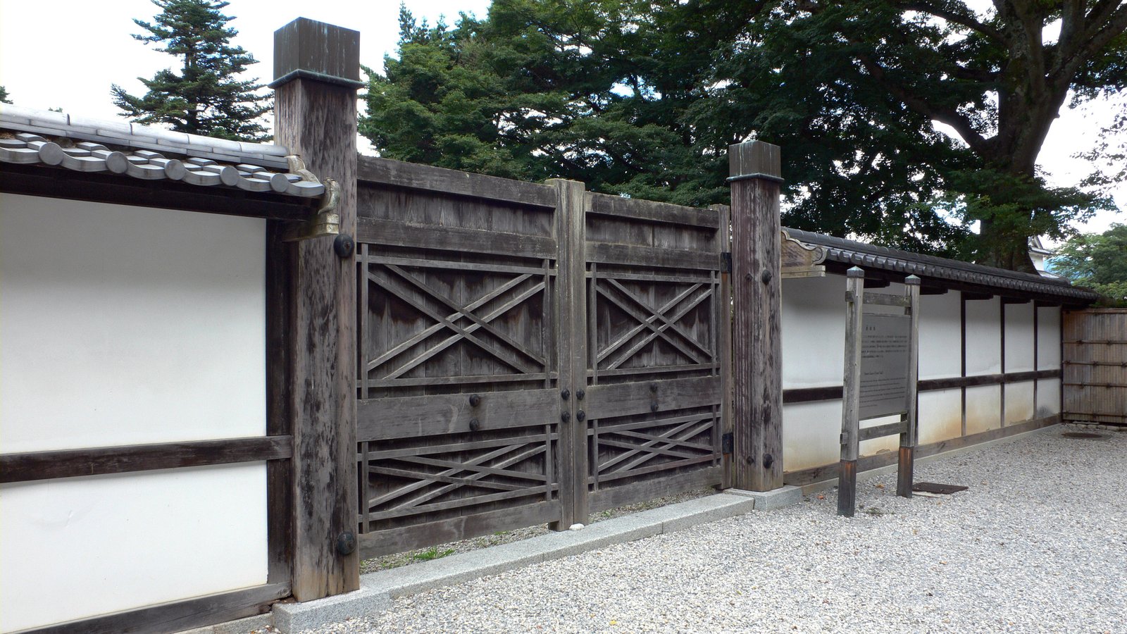 彦根城博物館の外まわり。白い塀の間に、木の桟を斜めに組んだ両開きの門が閉じていて、手前は砂利敷き