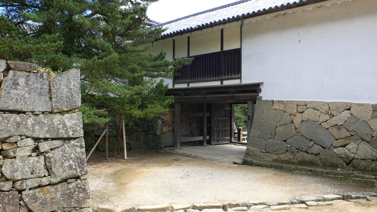 彦根城の天秤櫓。白漆喰の壁と黒い格子窓の櫓の下に木の門が開き、両側を灰色の石垣がはさむ。左手前に松の木