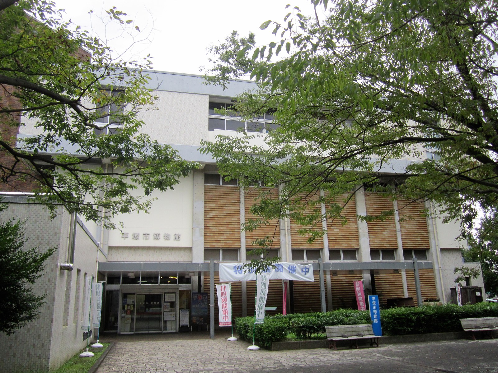 平塚市博物館の正面。「平塚市博物館」の館名を掲げた建物の入口に「特別展開催中」の横断幕とのぼりが立ち、手前に木々とベンチがある