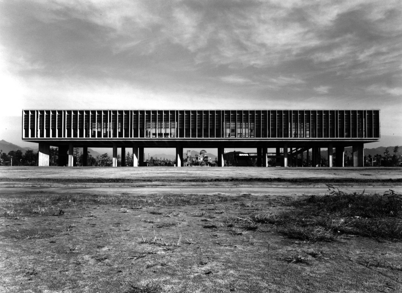 白黒写真。柱で持ち上げられた横長の建物が荒れ地の向こうに建ち、柱の間の奥に原爆ドームが小さく見える。空には厚い雲がかかっている