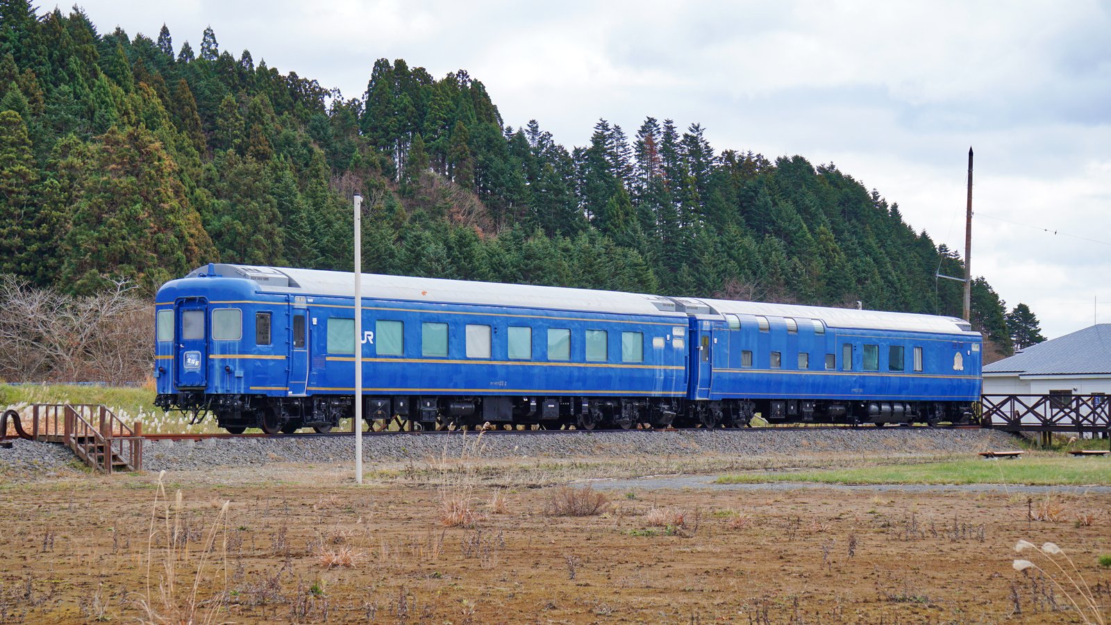杉林を背に短い線路の上に置かれた青い客車2両。車体に金色の帯とJRのマーク、先頭に北斗星のマークが付き、手前に木の階段がある