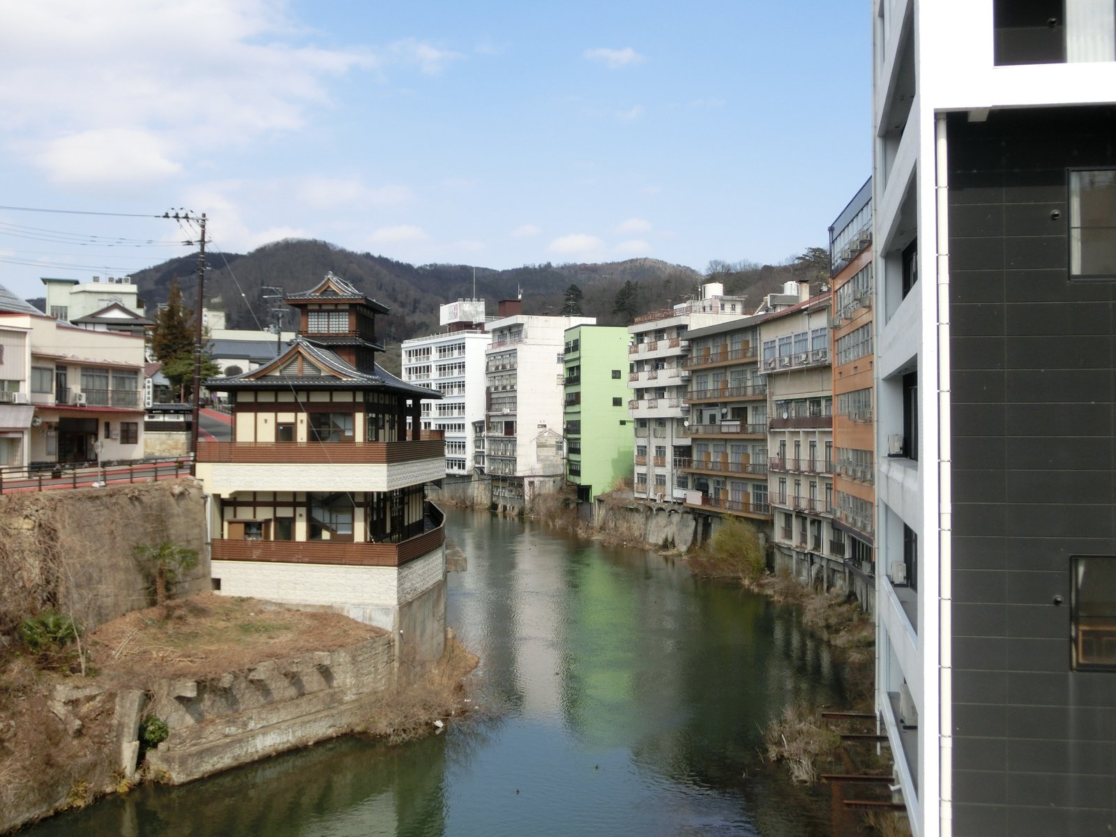 川の両岸に建物が並び、左岸には楼閣のような屋根を載せた和風の建物が見える。奥には低い山が連なる飯坂温泉