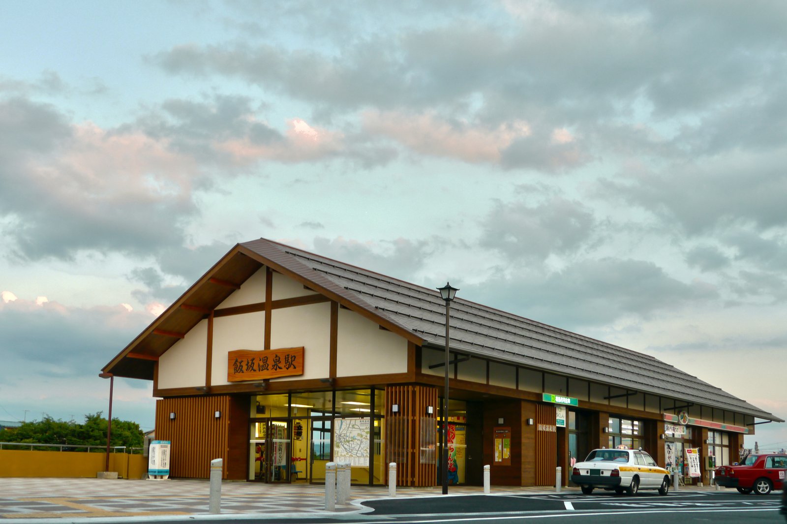 夕方の雲が広がる空の下、切妻屋根の和風の駅舎。破風の白壁に木の看板「飯坂温泉駅」が掛かり、手前の広場にタクシーが停まっている