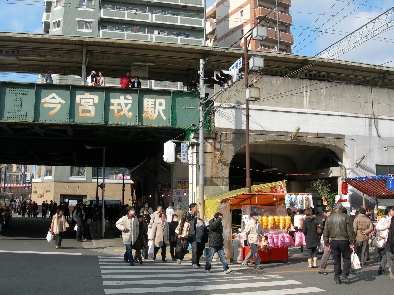高架の南海今宮戎駅。緑色の鉄桁に「今宮戎駅」の白い文字があり、下の道路を大勢の人が行き交い、右手の高架下に露店が出ている