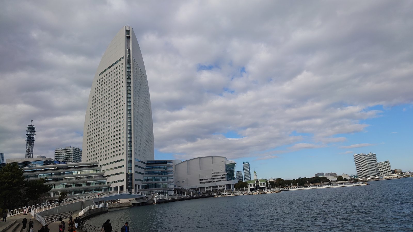 ヨコハマ グランド インターコンチネンタル ホテルを横浜港の岸から見たところ。半円形の白い高層棟が曇り空のもとに立ち、手前にパシフィコ横浜の低層棟、右に海が広がる