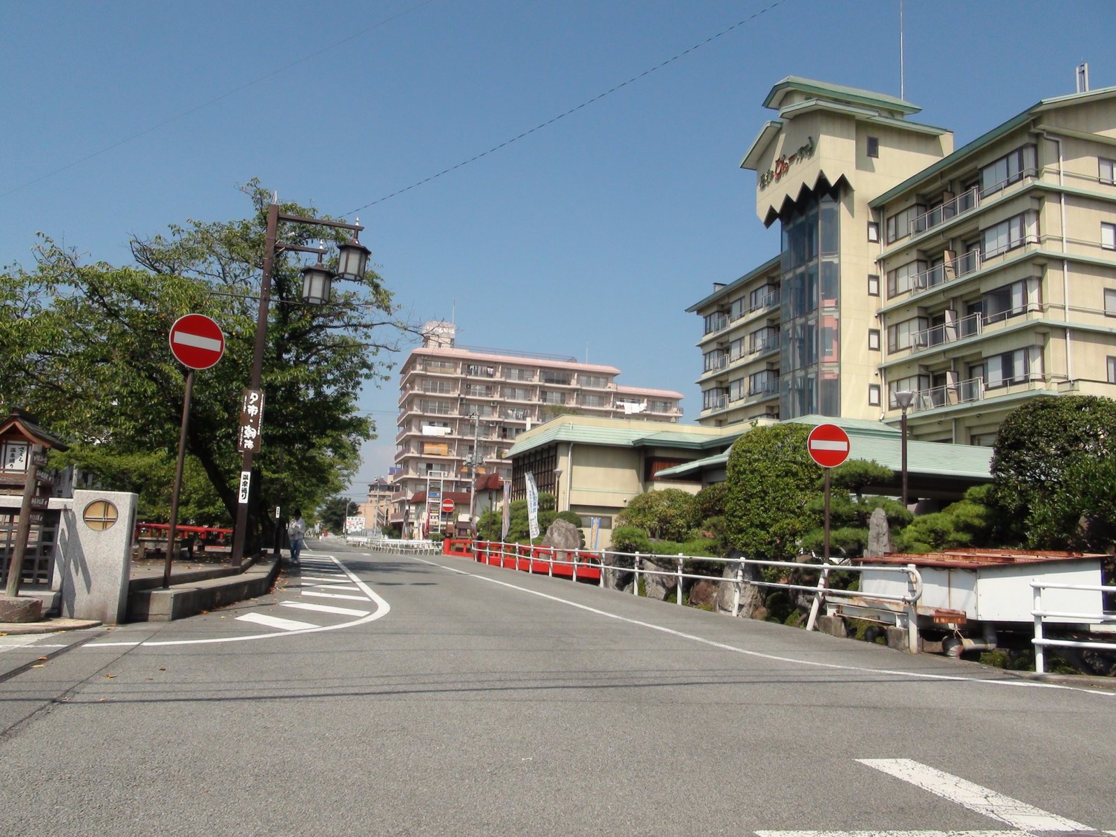 青空の下、石和温泉の道が緩く曲がる。右に屋根の瓦が緑色の大きな宿の建物と庭の松、奥に赤い欄干の橋と別のホテルが見える