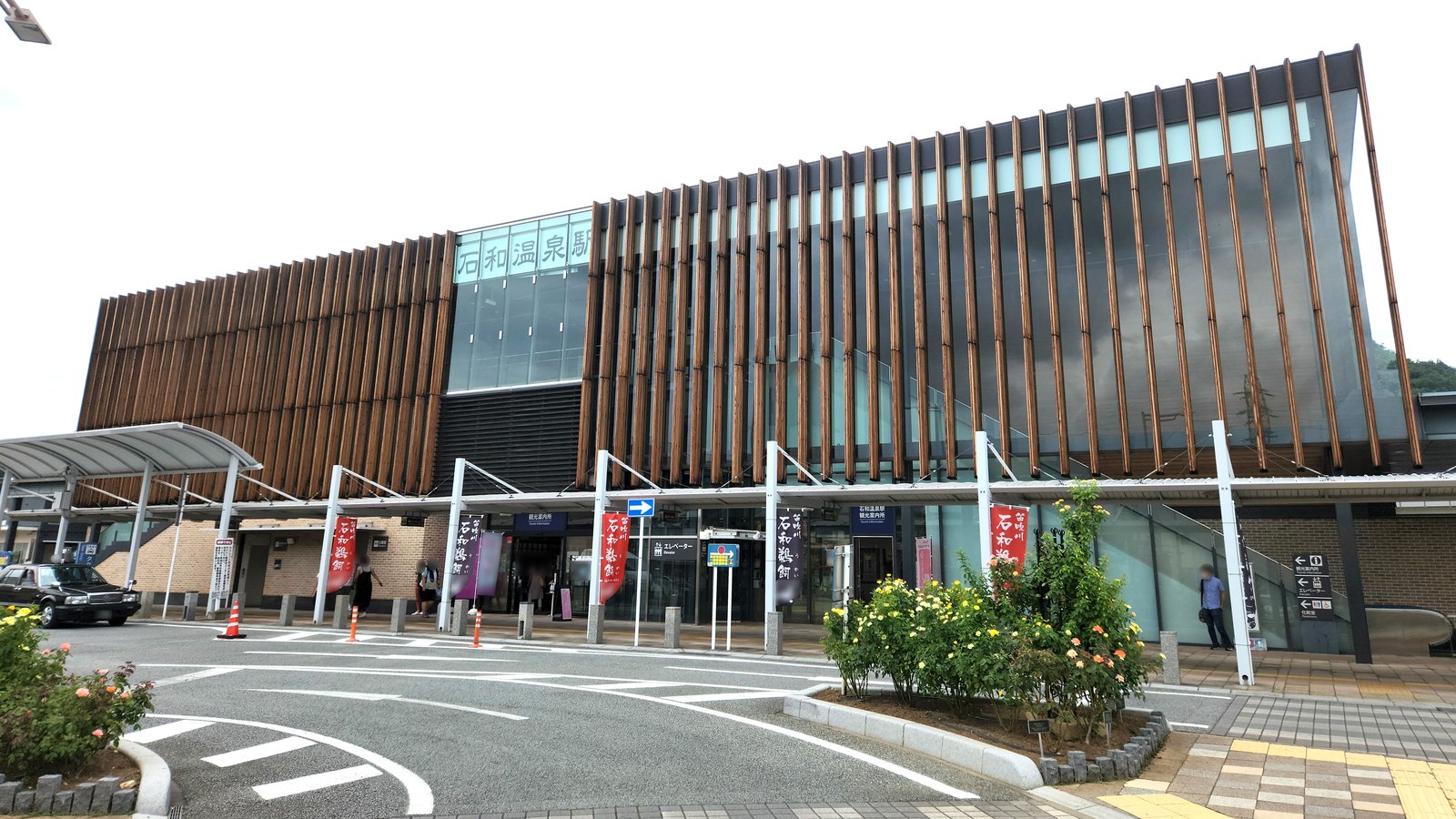 木の縦格子とガラスで覆われた石和温泉駅の南口駅舎。1 階に観光案内所の看板があり、手前にロータリーとバラの植え込みが見える