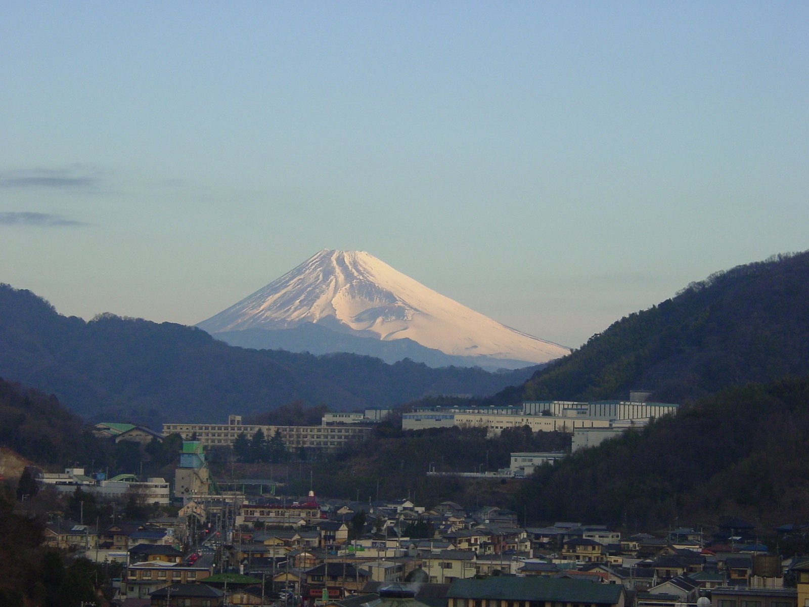 夕暮れの空の下、手前に伊豆長岡温泉の家並みが広がり、谷あいの山の間に雪をかぶった富士山の頂が薄紅色に染まって見える
