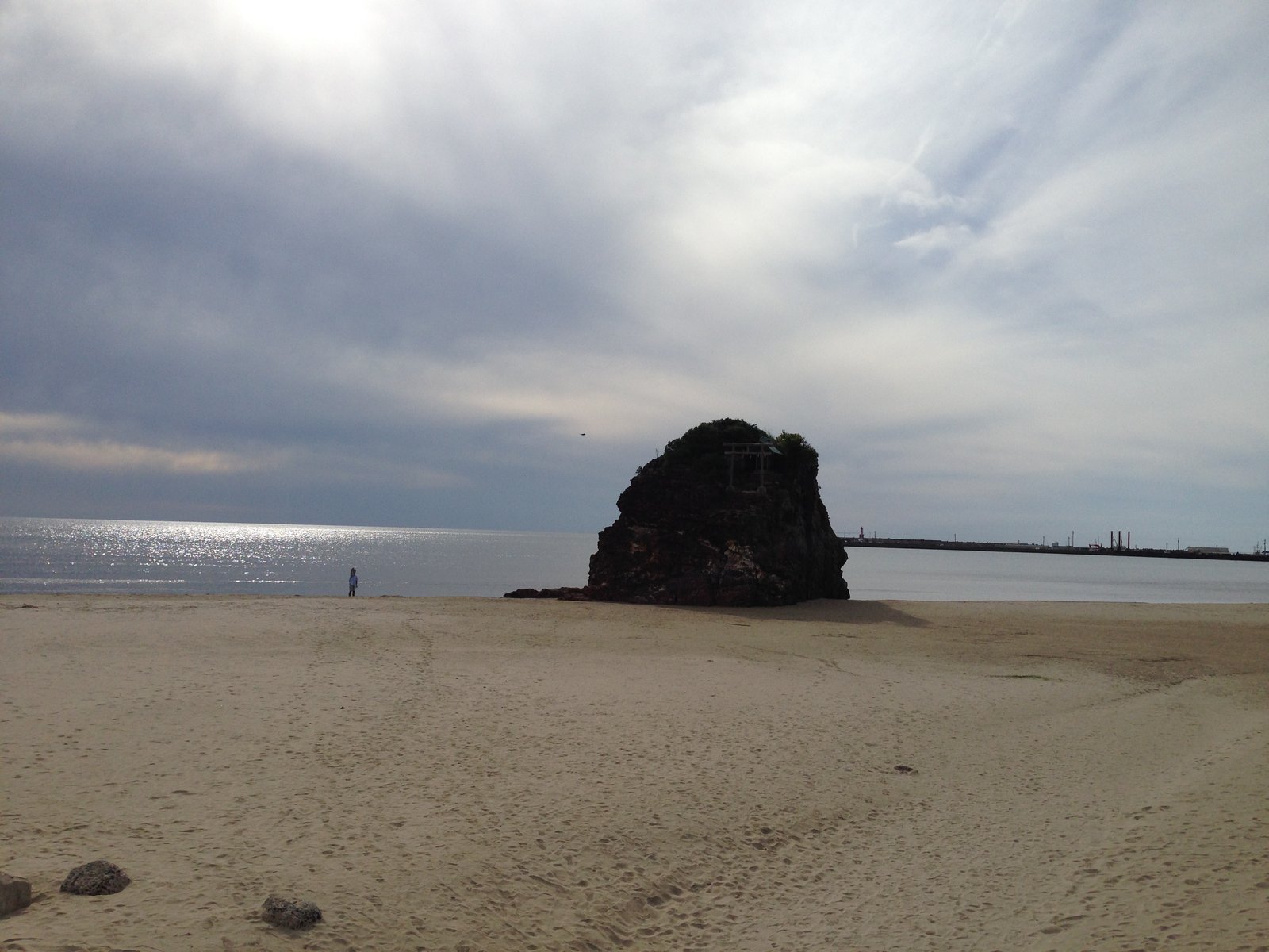 広い砂浜の先の海際に、頂に小さな鳥居と木のある黒い岩の島が立つ。空は薄い雲に覆われ、海面が光っている