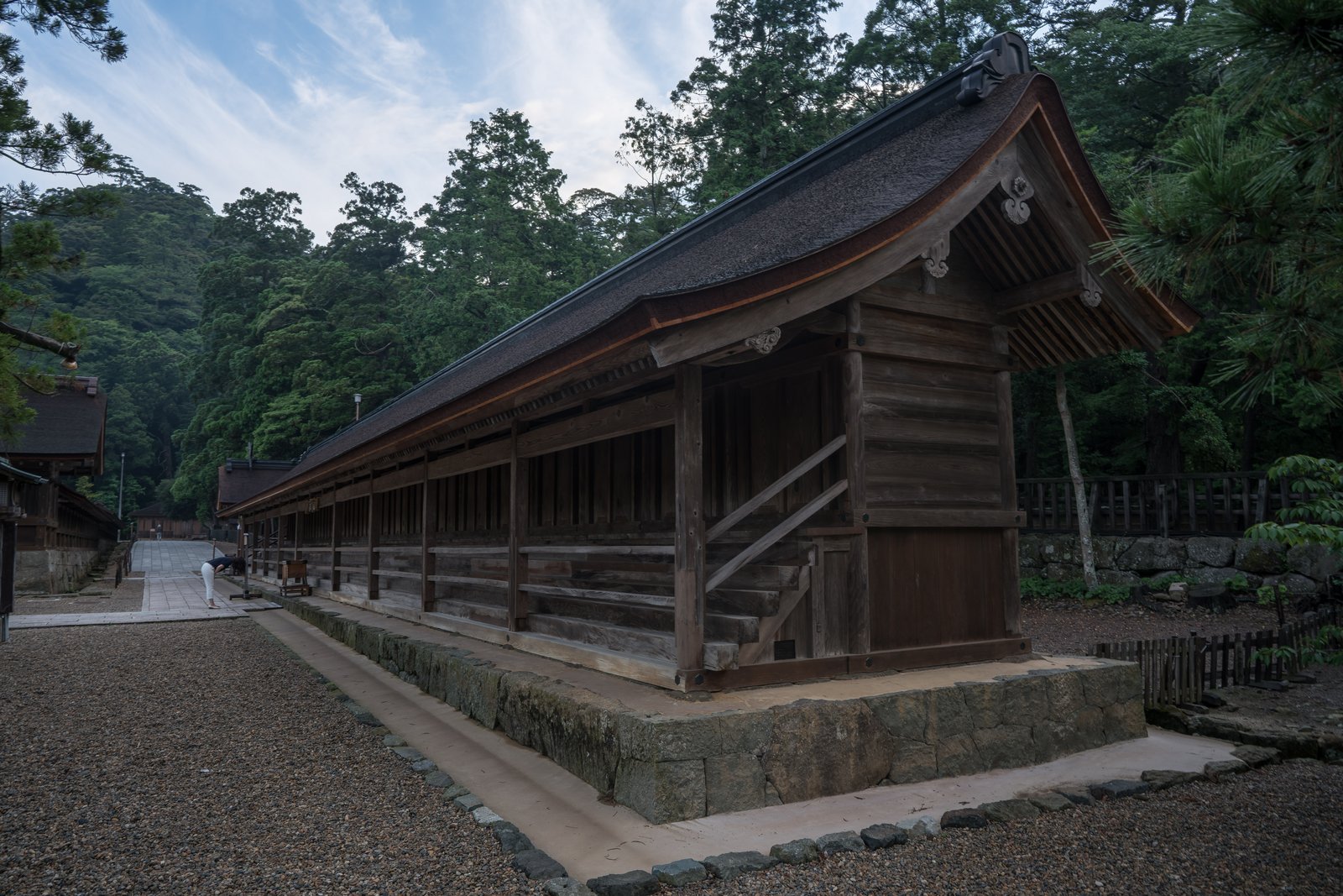 横に長い木造の社。反りのある屋根の下に格子の扉が連なり、石垣の基壇の上に建つ。奥に杉の木立と別の社殿