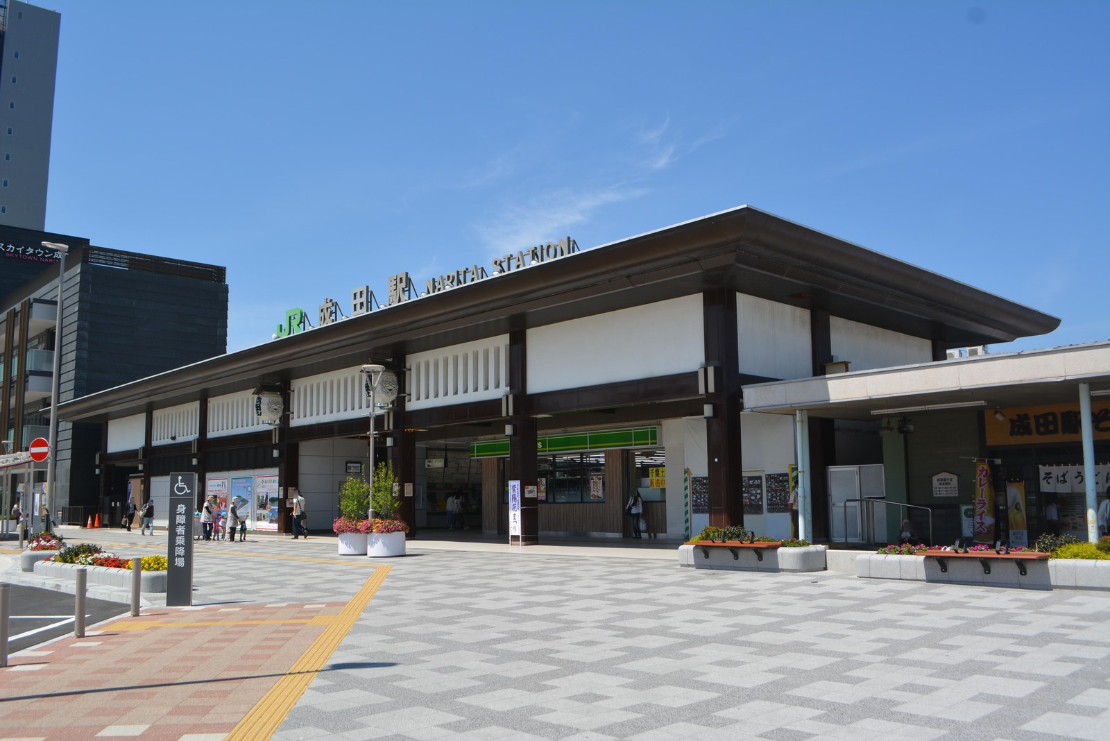 JR 成田駅の駅舎。瓦屋根をのせた平屋風の建物で、前に広場と花壇がある