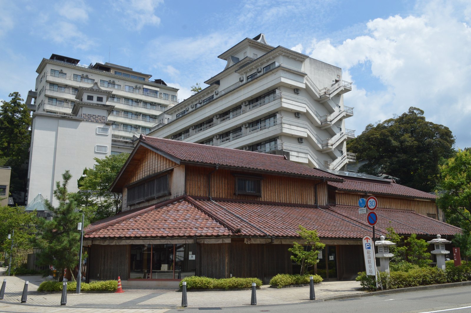 赤茶色の瓦屋根と木の壁の山代温泉総湯の建物。後ろに大きな旅館が2棟そびえ、手前に「総湯駐車場」の標識と石灯籠がある。