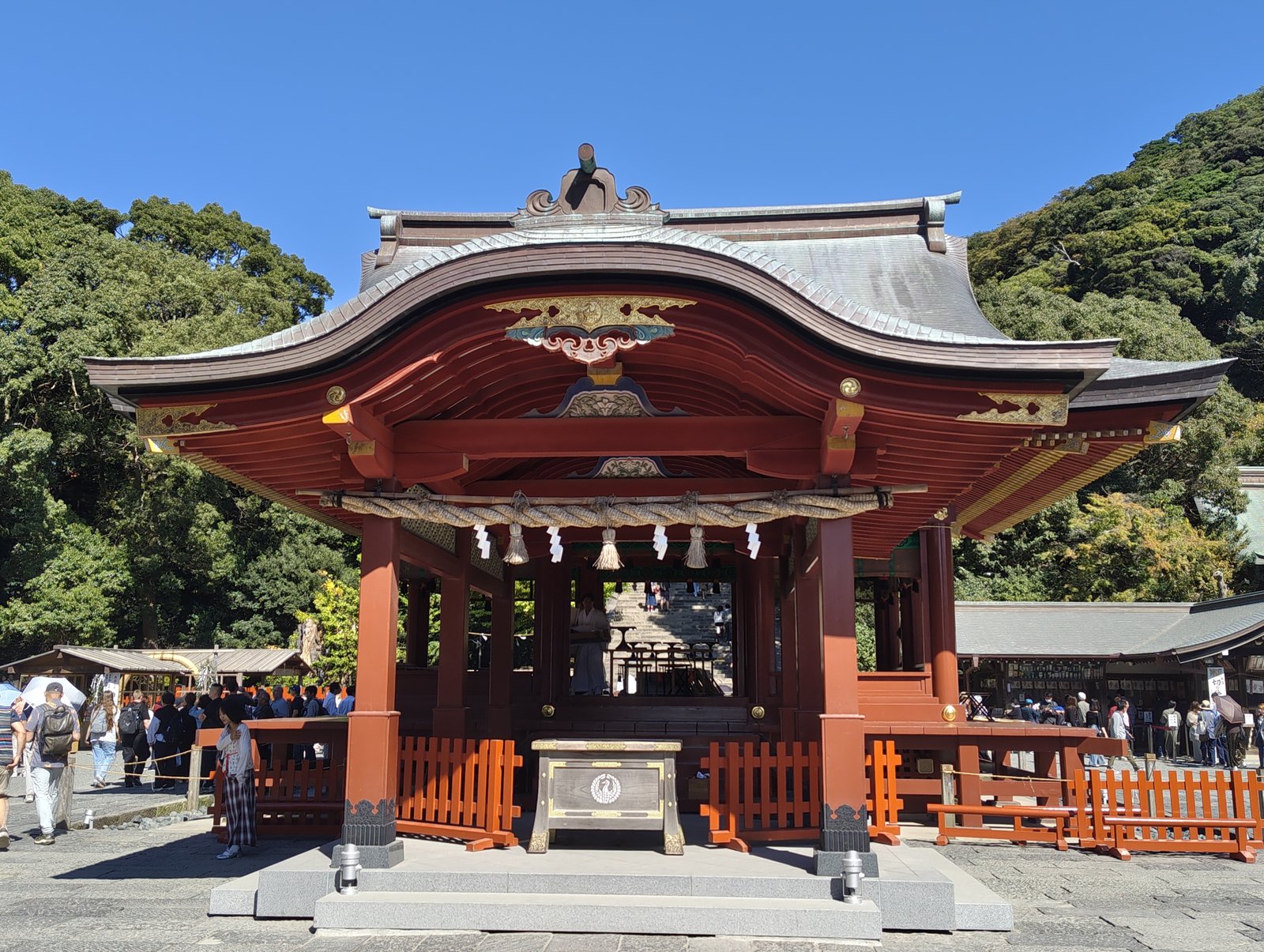 朱塗りの柱と反りのある屋根を持つ鶴岡八幡宮の舞殿。正面に注連縄と白い紙垂が掛かり、建物の奥に本宮へ上る大石段が見える。背後は木の茂る山で、空は青く、周りに参拝者が小さく写る