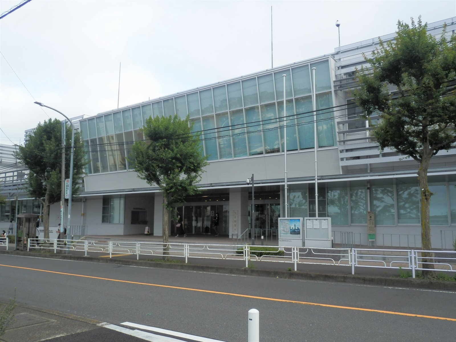 上の階がガラス張りになった白い横長の建物。手前に道路と街路樹がある