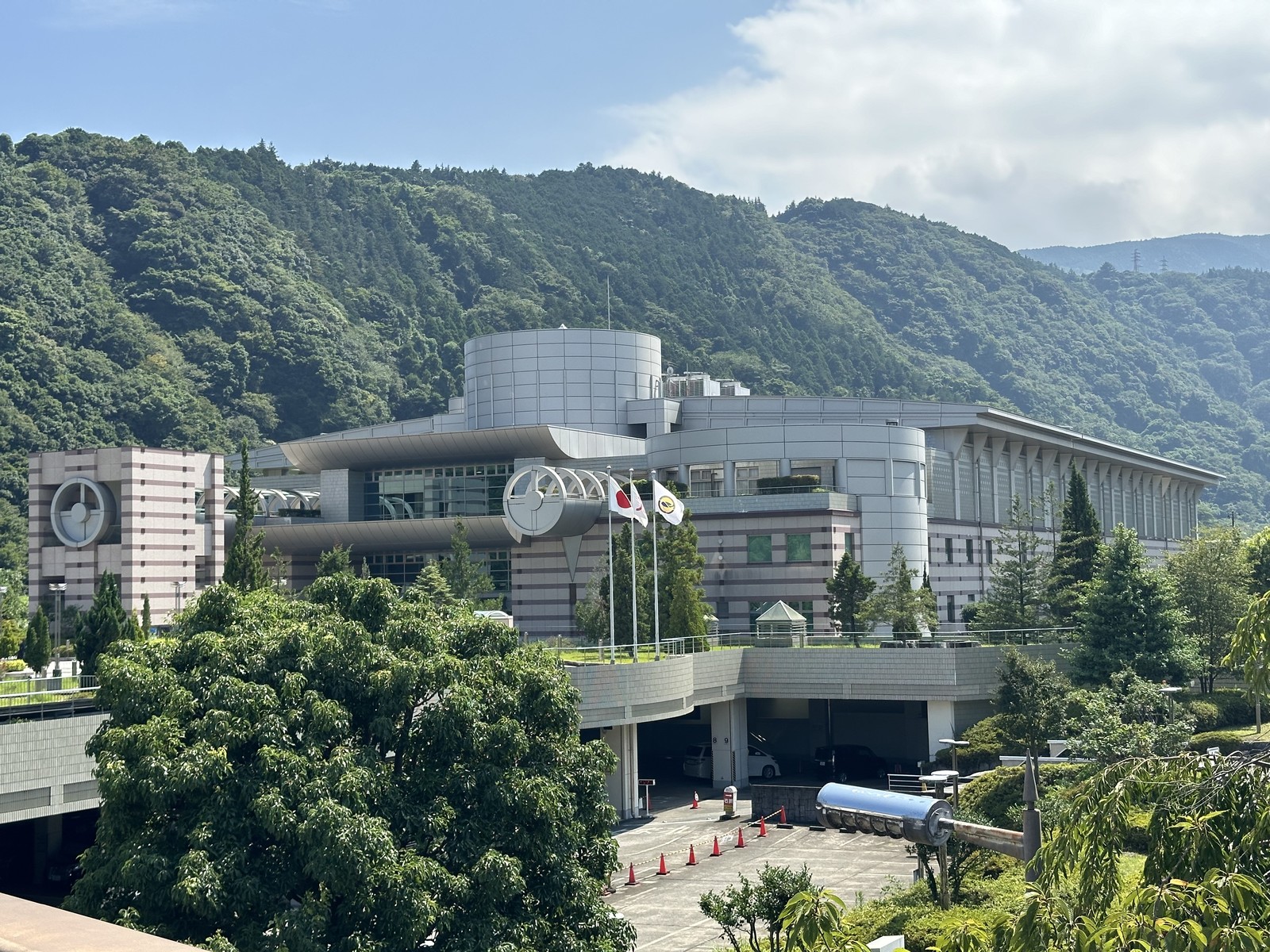 神奈川県立生命の星・地球博物館の外観。山を背に、円筒形の塔と丸窓を持つ灰色の建物が横に連なり、手前の広場に3本の旗竿と駐車場への下り坂
