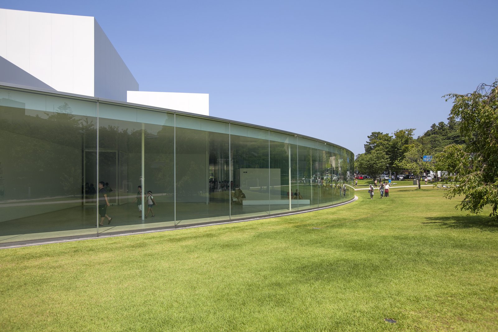 金沢21世紀美術館の円い建物の外壁。天井までの曲面ガラスが芝生に沿って続き、ガラス越しに館内を歩く人が見える。奥に白い四角い展示室が立ち上がる