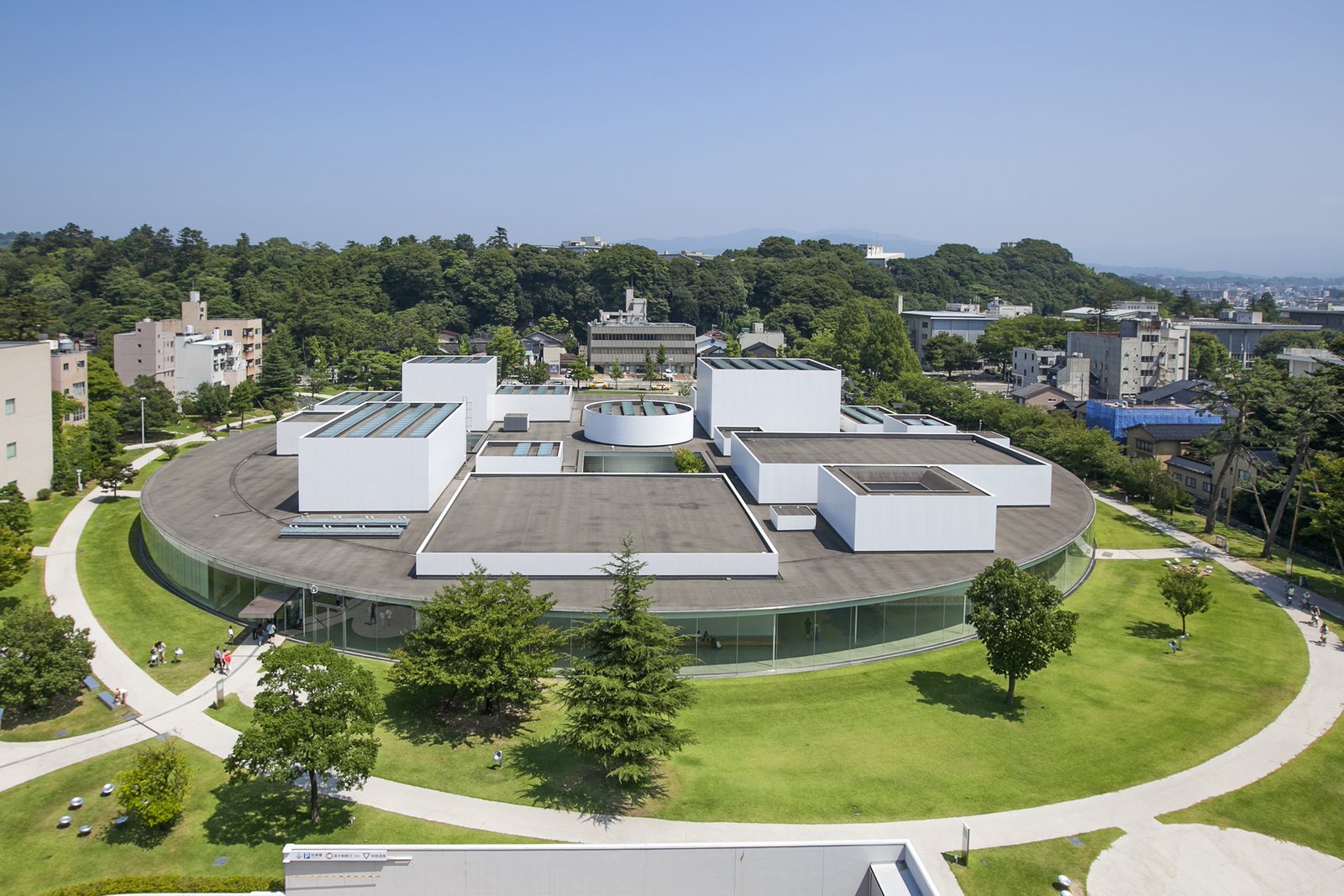 高い所から見下ろした金沢21世紀美術館。芝生と木に囲まれた円形の建物の平らな屋根から、大きさと高さの違う白い四角い展示室がいくつも突き出ている
