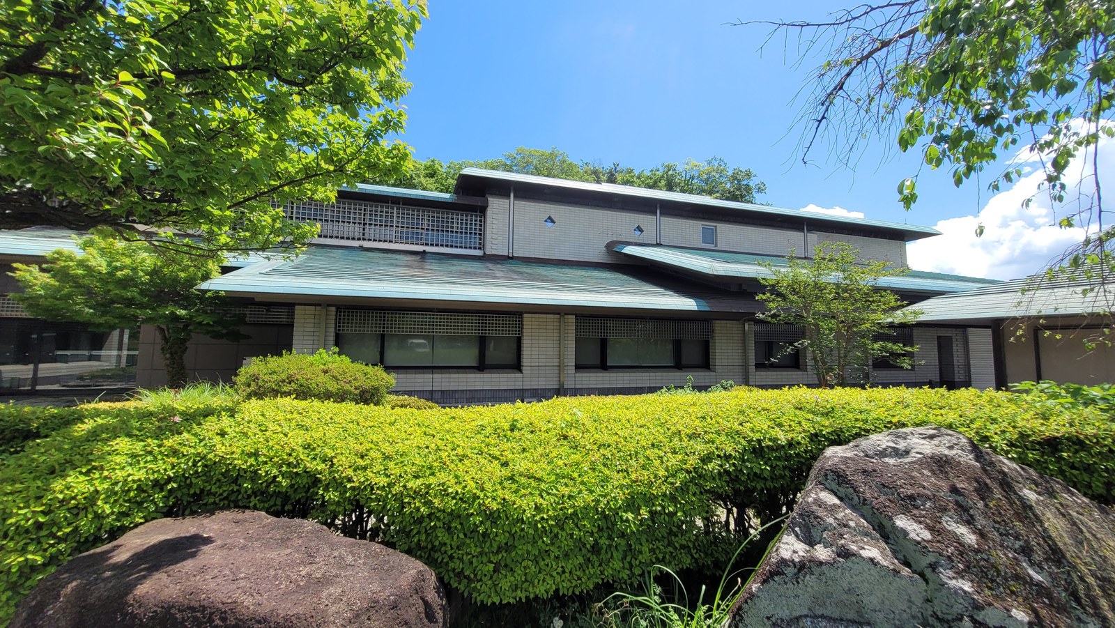 生垣と木立に囲まれた神奈川県立金沢文庫の建物外観。銅板葺きの大きな屋根と横長の窓が続く平屋づくり