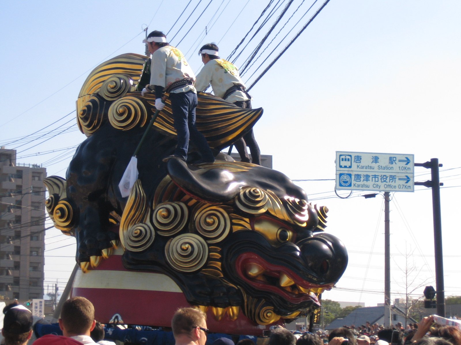 昼の通りを進む黒い獅子の曳山。金色の渦巻き模様のたてがみで、上に白い法被の人が 2 人乗る。右奥に「唐津駅 1.0km」の青い道路標識
