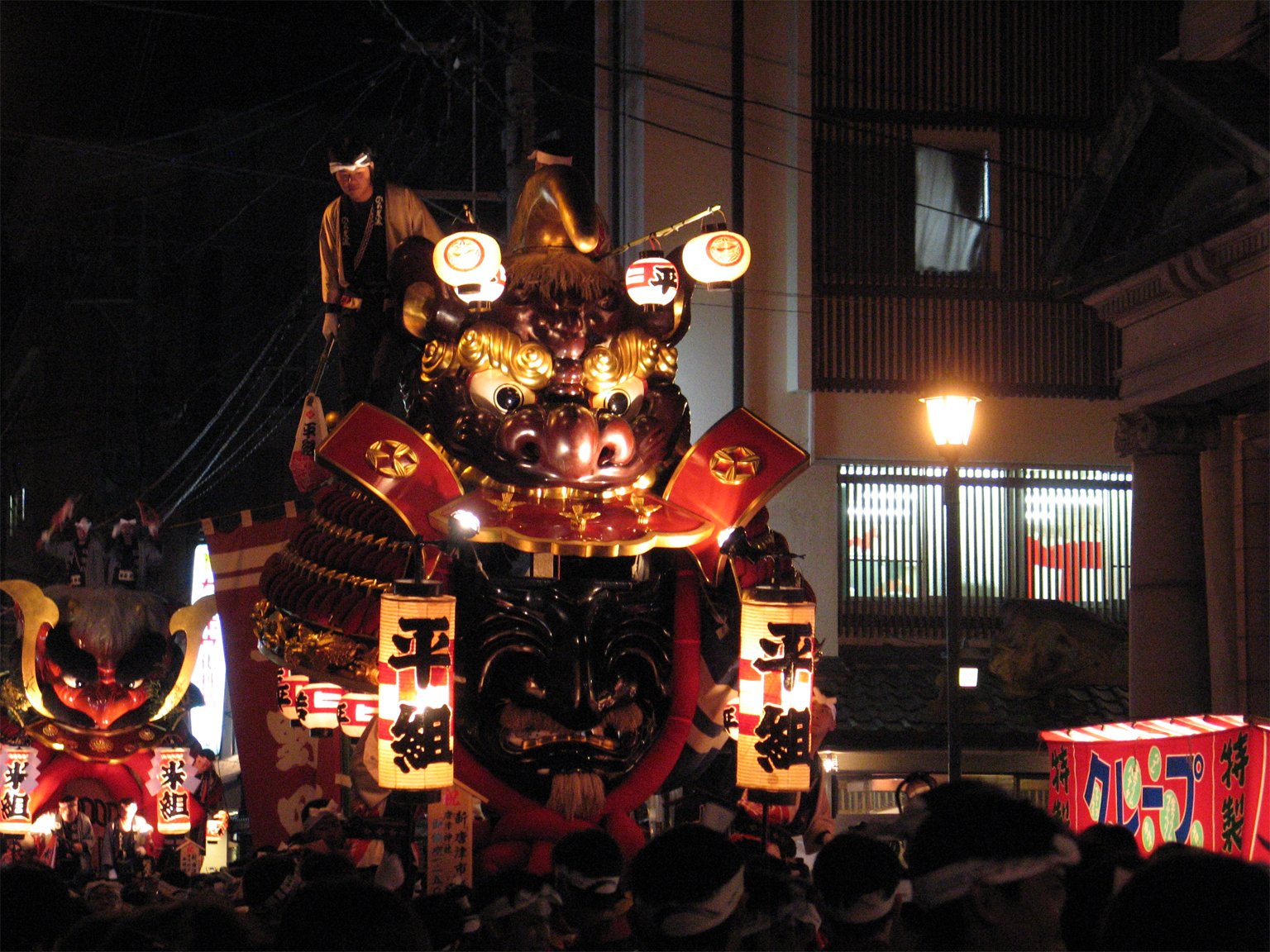 夜の通りを進む兜の形の曳山。黒い面と赤い鎧の飾りの上に法被の人が立ち、「平組」と書いた提灯が下がる。後ろにもう 1 台の曳山と見物の人の頭