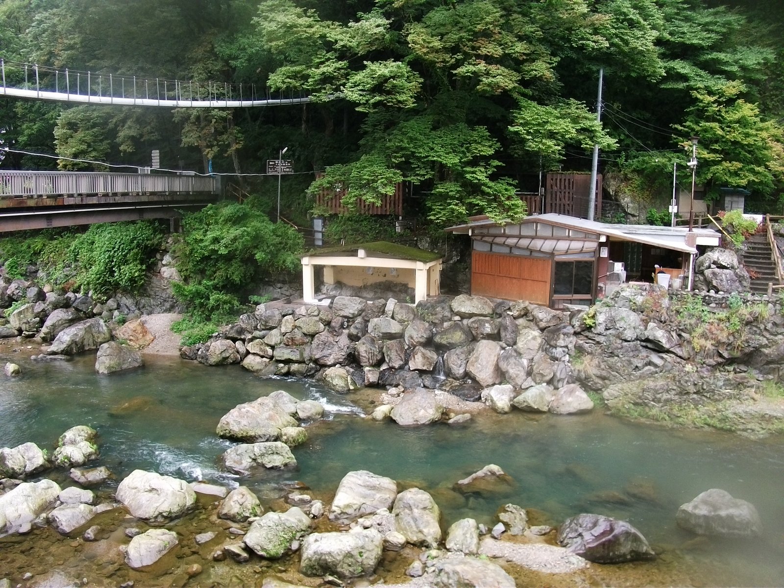 岩の多い川の岸に、苔の載った屋根の小さな湯小屋と木の壁の建物が並ぶ。上には吊り橋と道路の橋がかかり、背後は緑の木々に覆われている
